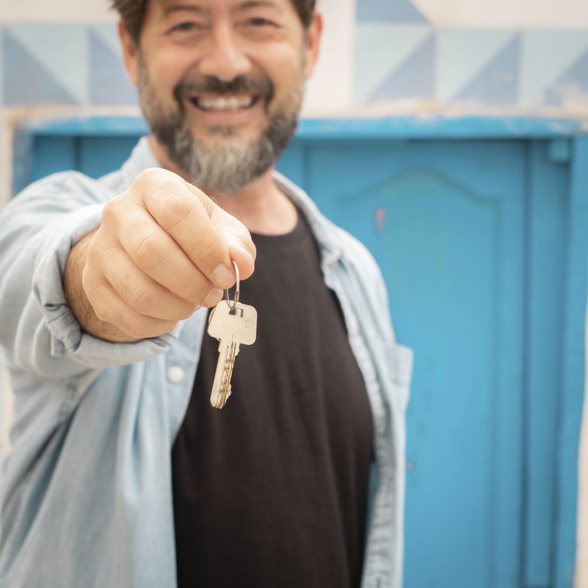 Adult happy man holding home keys and blue door house in background. Concept of buyer